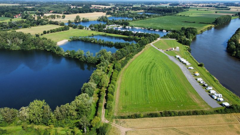 Landschaft mit Seen, Wiesen und einem Fluss, umgeben von Bäumen und Wohnmobilstellplätzen.